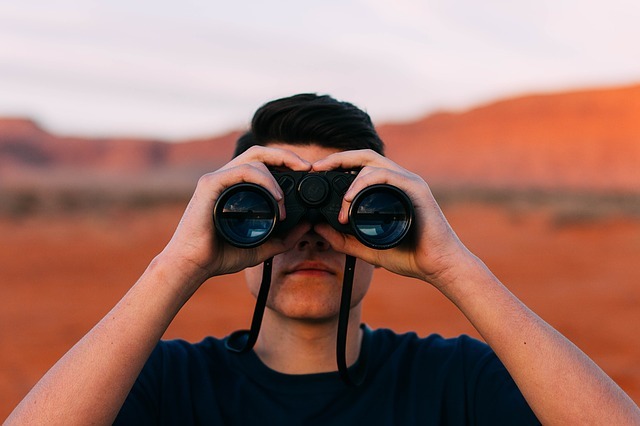 Photograph of a man searching through a pair of binoculars while outdoors.