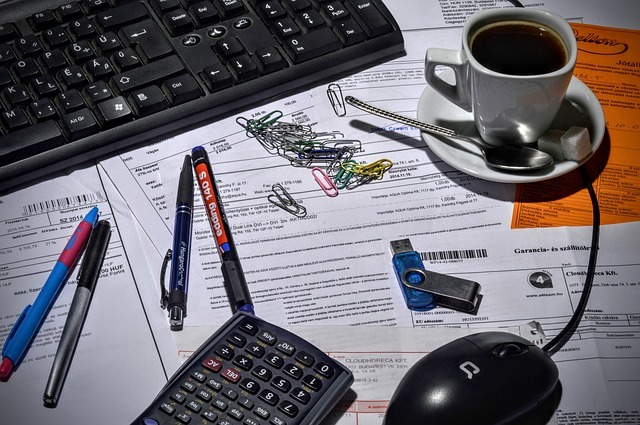 Photograph of invoices, paper clips, computer keyboard and mouse, and coffee sitting messily on a desk.