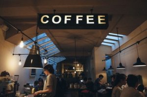 This is a photograph of a busy coffee shop with lots of people seated, including some on laptops. A sign hangs from the ceiling that says, "Coffee."