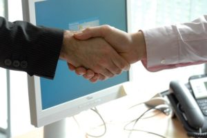 This is a photograph of two arms extended into a handshake in front of a computer screen.