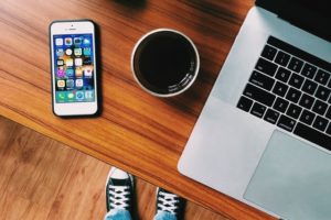 An above view photograph of a person's Converse sneaker-wearing feet on the floor beneath a wood desk where a phone, cup of black coffee, and a laptop sit.