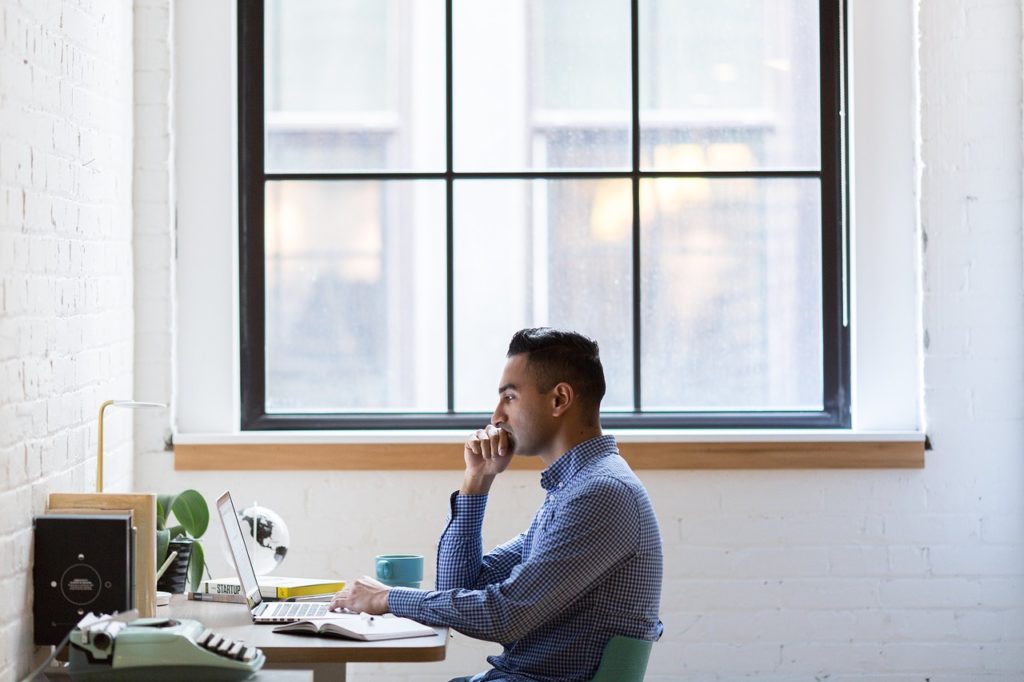 One of the rules of a redesign is to do your research. This image is a photograph of a man sitting at a computer and reading something on screen with an intense look on his face. There is a large window with no curtains beside him on the wall, letting a lot of natural light.