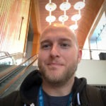 A photo head shot of Mike England, Frontend Engineer at WebDevStudios. he's slightly smiling at the camera while riding down an escalator and wearing a WordPress WordCamp lanyard.