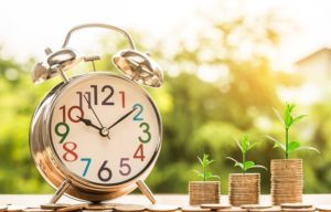 This is a photograph of a tabletop clock sitting next to three stacks of coins. From each stack of coins there is a green sprout growing.