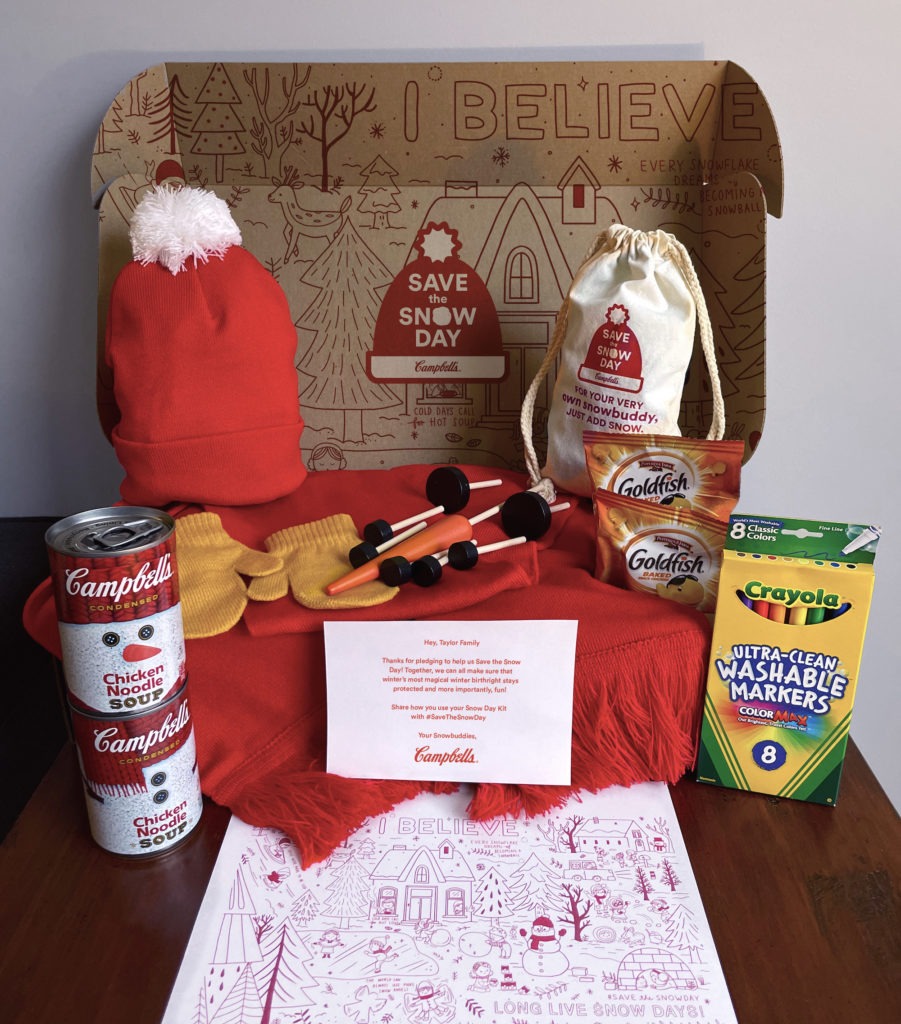 A photograph of the Campbell's Save the Snow Day Activity Kit, which includes cans of soup, a red knit cap, crayons, a coloring sheet, mittens, and accessories for building a snowman.