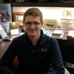 A portrait of Backend Engineer for the website design and development agency WebDevStudios, Scott Anderson. Scott is sitting at a desk, looking at the camera, and smiling.